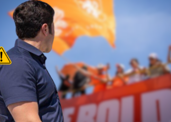 Man Seen from Behind at a Rally, Facing a Cheering Crowd with an Orange Banner and a Warning Icon Overlaid on the Left Side. - Regio Politica