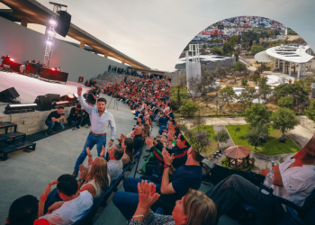 Male Performer on a Stage Waving to a Large Seated Audience at an Outdoor Concert, Sunset Sky Above the Venue. - Regio Politica