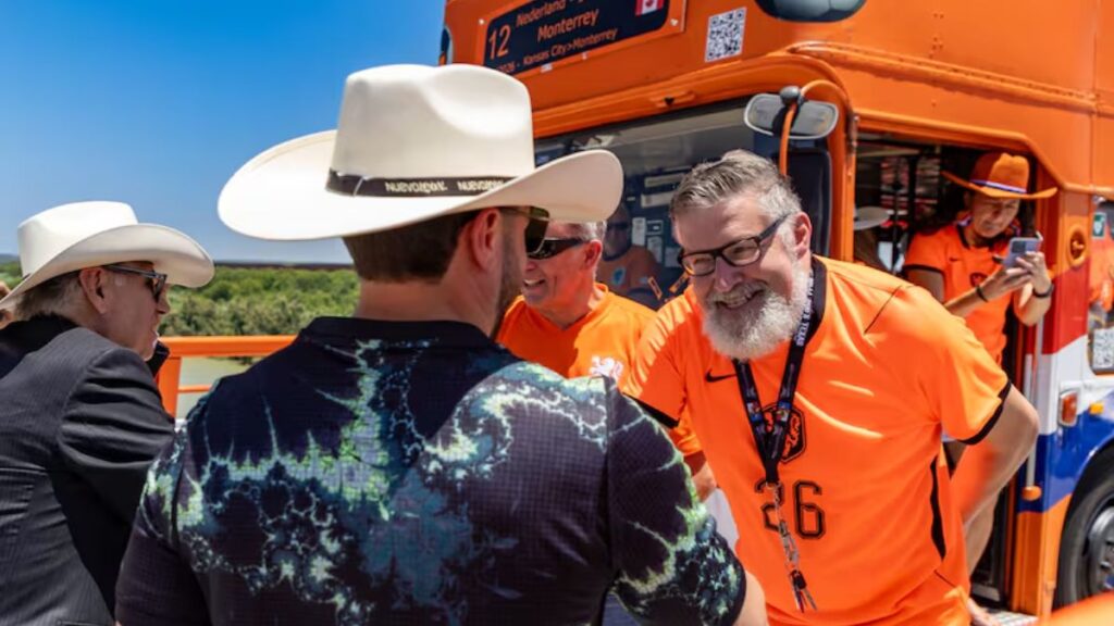 Smiling Man in Orange Jersey with Number 26 Greets Another Man Near an Orange Bus Steadied by a Group of People in Hats and Polo Shirts - Regio Politica
